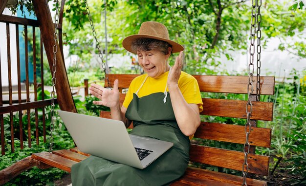 Senior woman gardener in a hat is watching something on a laptop computer and making a video call in the yard while outdoors