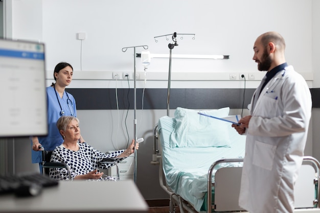 Senior woman getting medicine through intravenous line bag sitting on wheechair pushed by nurse in hospital room. Medical physician ward asking about pain to set diagnosis.