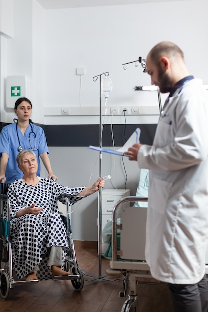 Senior woman getting medicine through intravenous line bag sitting on wheechair pushed by nurse in hospital room