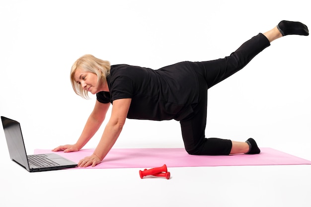 Senior woman goes in for sports at home on the mat in front of the laptop