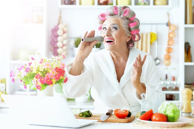 Senior woman in  hair rollers at kitchen with laptop and vegetables