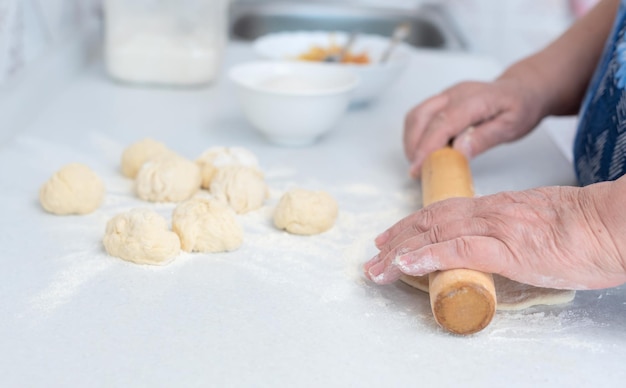 Senior woman hands rolling out the dough with a rolling pin on a white kitchen table with blurred grated apple and sugar on background Selective focus Process of making pies with apple filling