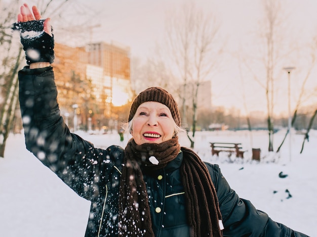 senior woman in hat and sporty jacket snowballing in snow winter park