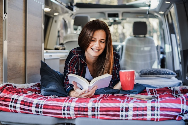 Senior woman having inside camper van reading a book and drinking coffee - Focus on face