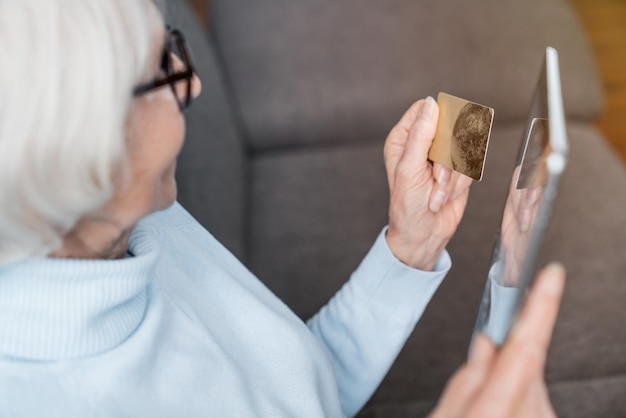 Senior woman holding bank card while shopping online on digital tablet sitting on sofa at home