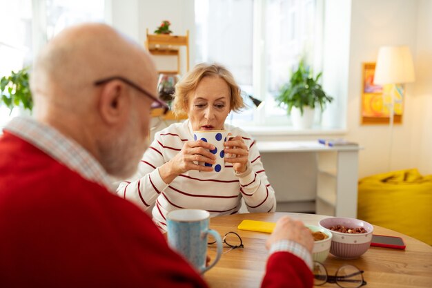 テーブルで彼女の友人の向かいに座っている間おいしいお茶とカップを保持している年配の女性