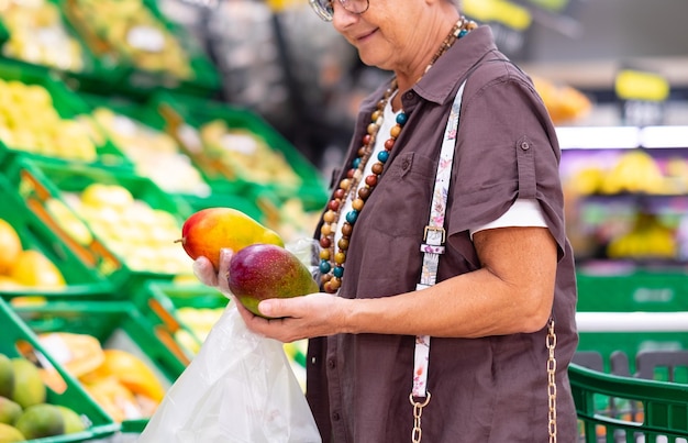 スーパー マーケットや食料品店のクローズ アップで 2 つの新鮮なマンゴーを保持している年配の女性 女性は保護手袋を着用して 2 つの成熟したマンゴーを保持しています