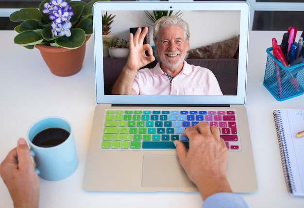 A senior woman holds up a cup of coffee during a videocall with her smiling husband - two retirees stay in touch remotely. White desktop outdoors on the terrace