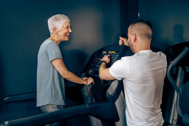 Senior woman is exercising on a treadmill with coach in the gym.