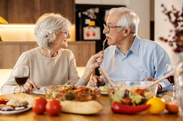 한 노인 여성 이 집 의 점심 테이블 에서 남편 에게 먹이 를 주고 있다