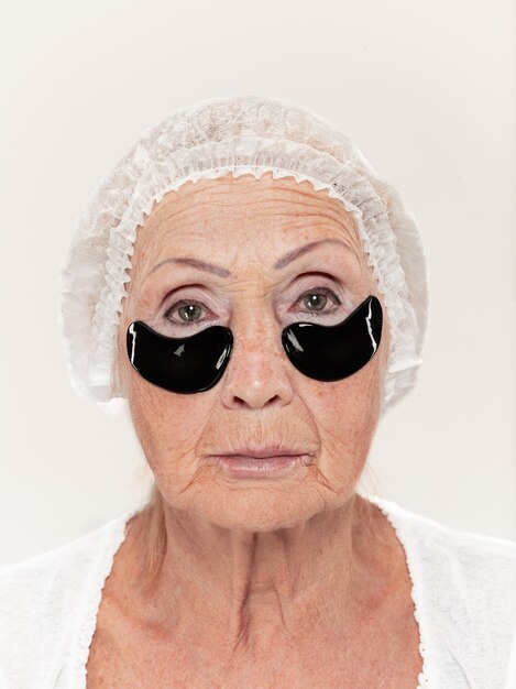 Senior woman at isolated on white wall looking at camera and making masks under eyes