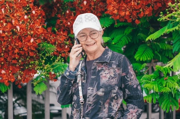 Photo senior woman in jacket jeans and white cap walking in spring park outdoors using mobile phone