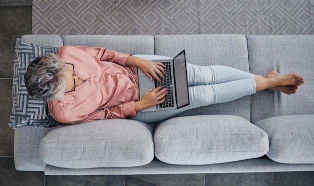 Senior woman laptop and sofa above relaxed in the living room checking email typing or writing at home Elderly female freelancer or writer relaxing on lounge couch working or reading on computer