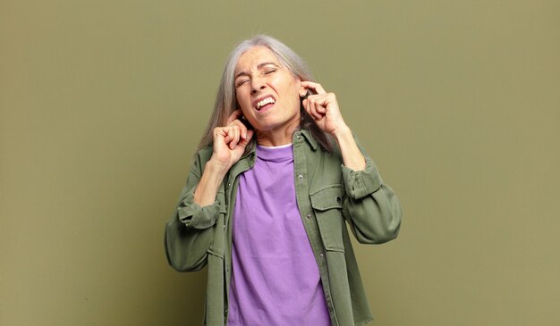 Senior woman looking angry, stressed and annoyed, covering both ears to a deafening noise, sound or loud music