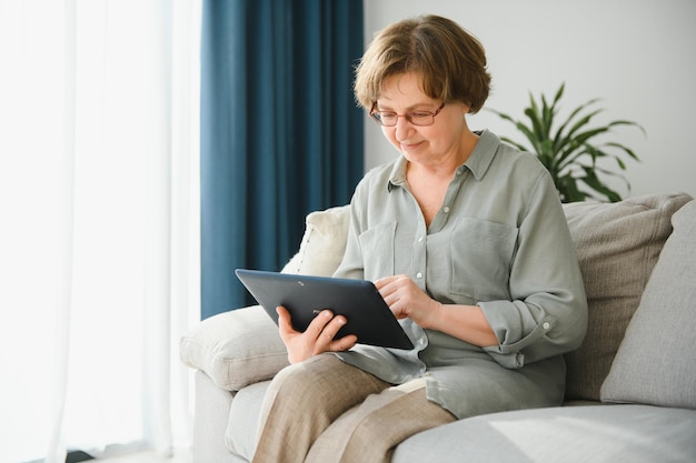 소파에 있는 디지털 태블릿을 보고 웃고 있는 노인 여성