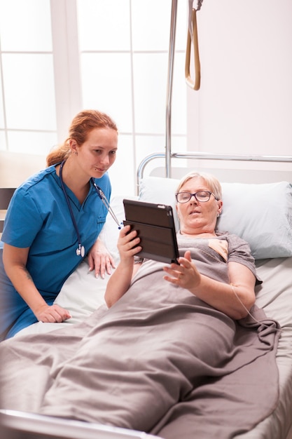 Старшая женщина, лежа в кровати дома престарелых, показывая что-то женской медсестре в планшетном компьютере.