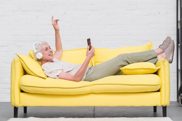 写真 ヘッドホンを介してモバイルで音楽を楽しむ黄色のソファに横たわっている高齢の女性