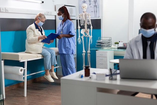 Senior woman patient with protective face mask signing medical documents during medical appointment in hospital office. Therapist nurse explaining sickness diagnosis discussing medication treatment