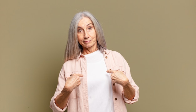 Senior woman pointing to self with a confused and quizzical look, shocked and surprised to be chosen