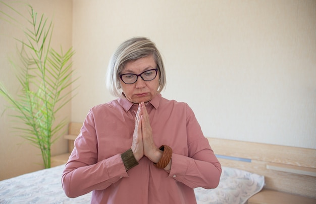 Senior woman prays to God for loneliness in her home in bedroom. Concept psychological health emotional anxiety
