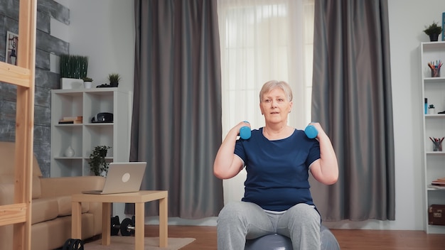 Senior donna di pompaggio del muscolo della spalla utilizzando manubri seduto sulla sfera di equilibrio. la persona anziana che si allena a casa pratica uno stile di vita sano, allenamento per anziani fitness in appartamento, attività e assistenza sanitaria