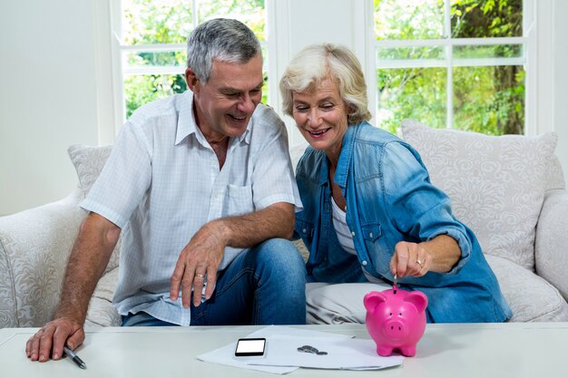 Senior woman putting coins in piggy bank while sitting with man on sofa