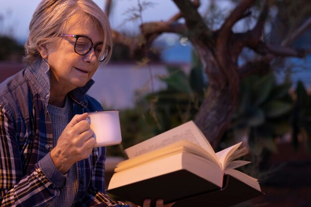 Foto donna anziana che legge un libro mentre tiene il caffè all'aperto