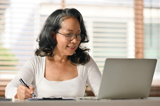 Senior woman reading online news browsing internet on laptop at home Retired lifestyle and technology concept