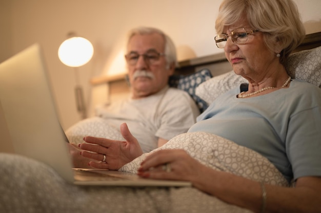 Senior woman reading problematic email on laptop while lying down in bed with her husband at night