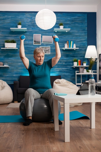 Старшая женщина на пенсии поднимает руки, делает упражнения с гантелями, сидя на фитболе ...