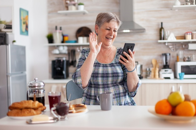Senior woman saying hello while having video call with family using smartphone in kitchen during breakfast. Elderly person using internet online chat tech, tablet webcam for virtual conference call