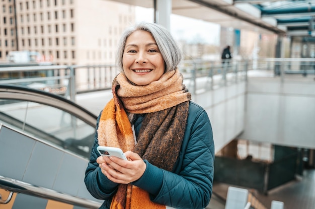 La donna senior invia un messaggio con un telefono cellulare in una stazione di trasporto