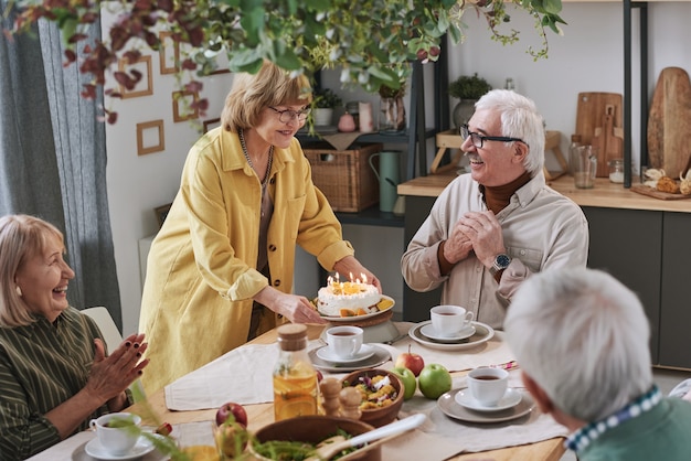 Senior woman serving birthday cake for senior man while they sitting at the table with friends at home