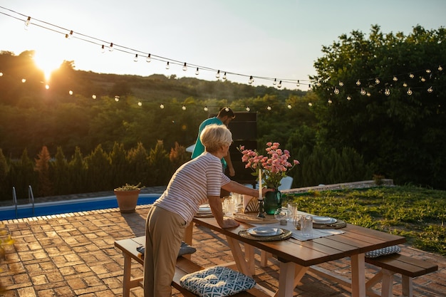 Senior woman setting dining table in the backyard and preparing everything for family gathering