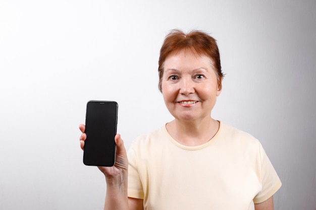 La donna senior mostra un telefono su bianco in una maglietta leggera