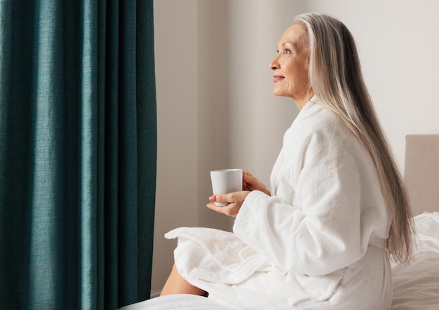 Senior woman sitting on bed in bedroom and looking away Side view of pensive mature femalex9xA