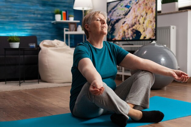 Senior woman sitting comfortable in lotus position on yoga mat with closed eyes meditating