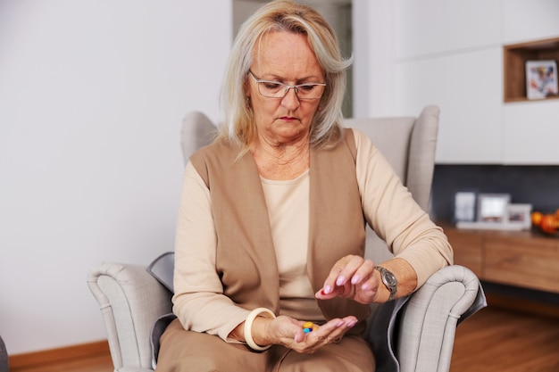 Senior woman sitting at home in her chair and holding hand full of pills and vitamins