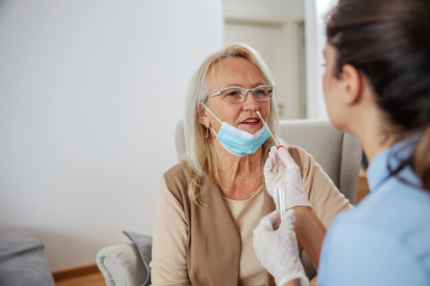 Senior donna seduta a casa mentre infermiera utilizzando un tampone di cotone durante l'epidemia di virus corona.