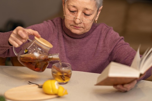 Senior woman sitting at the table drinking tea with lemon and reading a book