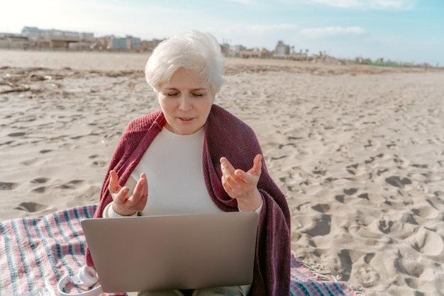 Senior woman sitting with her laptop on the beach and talking to someone during the video call