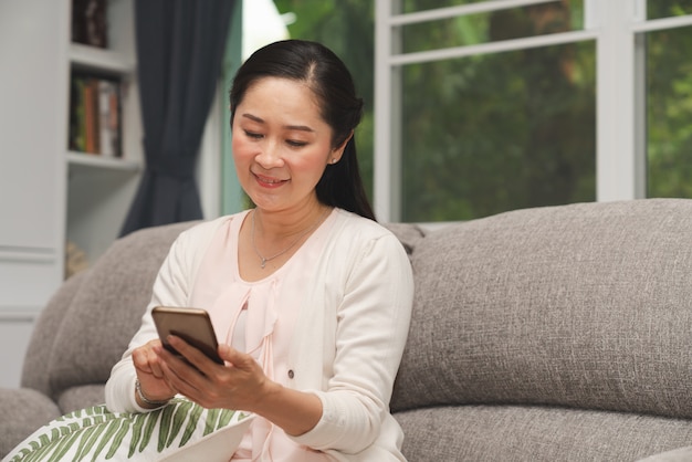 年配の女性が自宅の居間のソファーにスマートフォンを見ながら笑顔します。