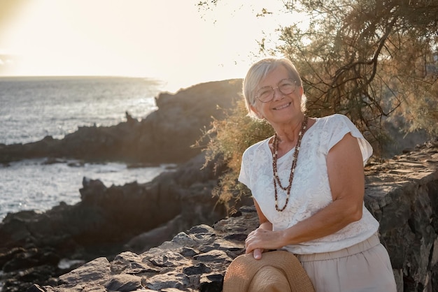 Senior woman standing close to the sea at sunset looking at camera enjoying summer vacation