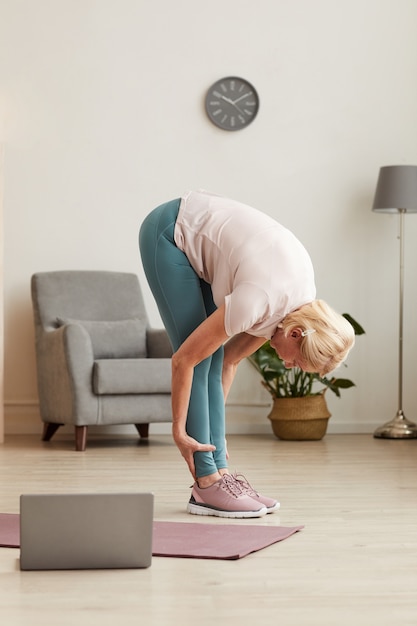 Senior woman standing and stretching her body during sports training in the room at home