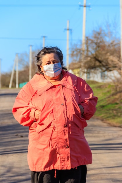 Senior woman stands in a medical mask on her face, which protects against coronavirus and other viruses and diseases on a rural street. Coronavirus pandemic