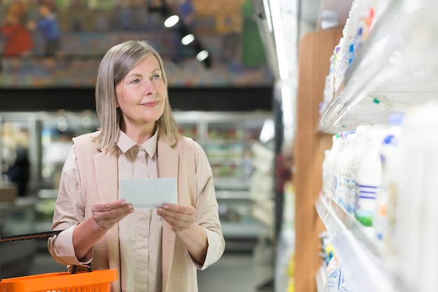 Senior woman in the store chooses goods in the dairy department near the refrigerators