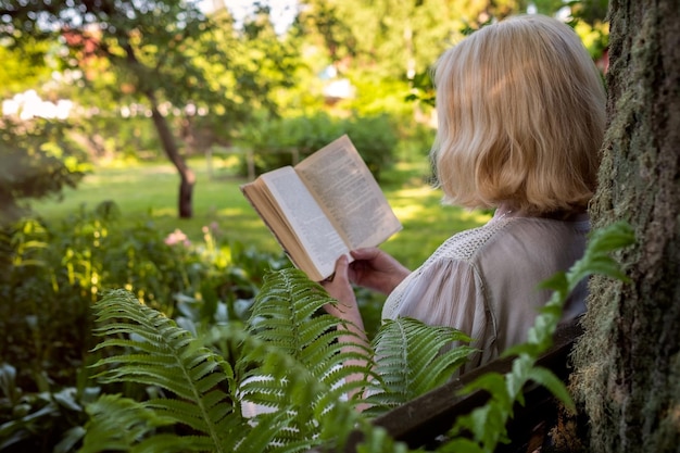 Пожилая женщина в летнем саду читает книгу, сидя возле дерева