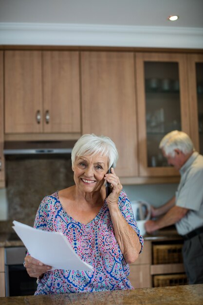 Senior woman talking on mobile phone while man working in kitchen at home