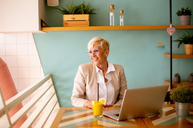 Senior woman using mobile phone while working on the laptop and drinking fresh orange juice in the cafe