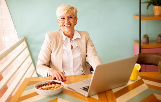Пожилая женщина пользуется мобильным телефоном во время работы на ноутбуке и завтракает в кафе